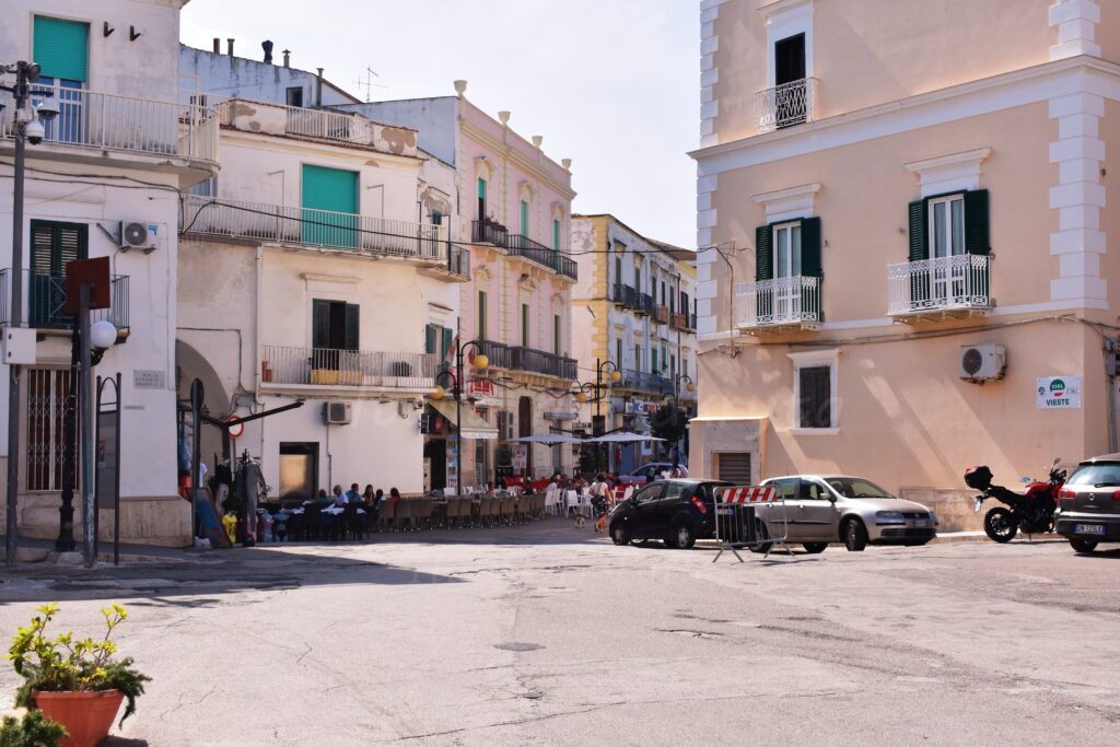 A sunny town square in Vieste, Puglia, surrounded by multi-story white and pastel-colored buildings with balconies, outdoor cafe seating, and parked cars and motorcycles