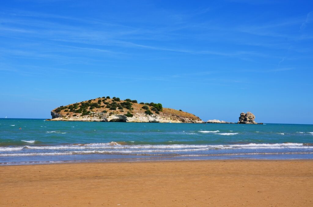 Sandy shore of Scialmarino beach in Vieste with view of the small, rocky Isola di Chianca covered in green shrubs under a bright blue sky, Gargano, Puglia