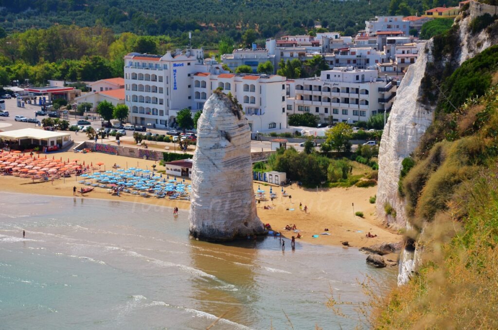 High-angle view of the massive Pizzomunno white limestone monolith on a sandy beach in Vieste, with beach umbrellas, coastal hotels, and white cliffs in the background, Gargano, Puglia.