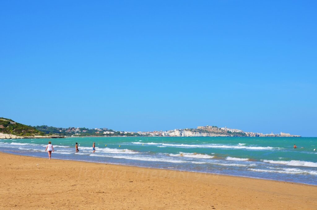 Wide sandy beach near Vieste with people wading in the turquoise Adriatic Sea and the town skyline on white cliffs in the background, Gargano, Puglia