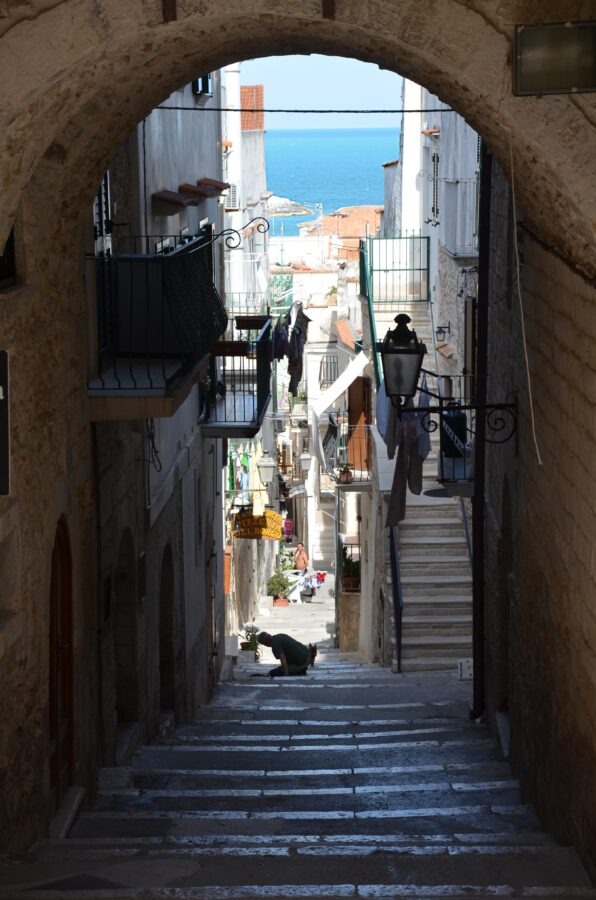 View through a dark stone archway looking down a narrow, steep alley with stone stairs in the old town of Vieste, leading toward the blue sea visible in the background