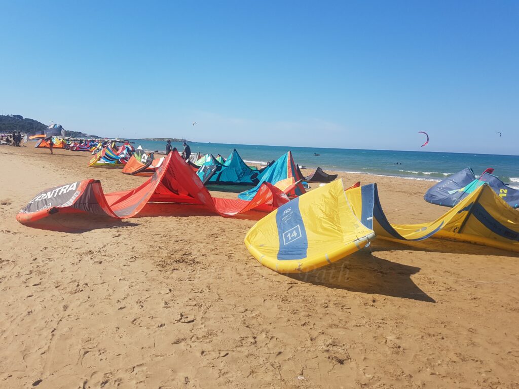 Brightly colored kitesurfing sails, including red, yellow, and blue ones, resting on a wide sandy beach next to the sea under a clear blue sky, Vieste, Gargano, Puglia.
