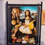 Street art z parodią Mony Lisy w fartuszku, otoczoną włoskimi produktami spożywczymi – wędlinami, serami, warzywami i rybą.