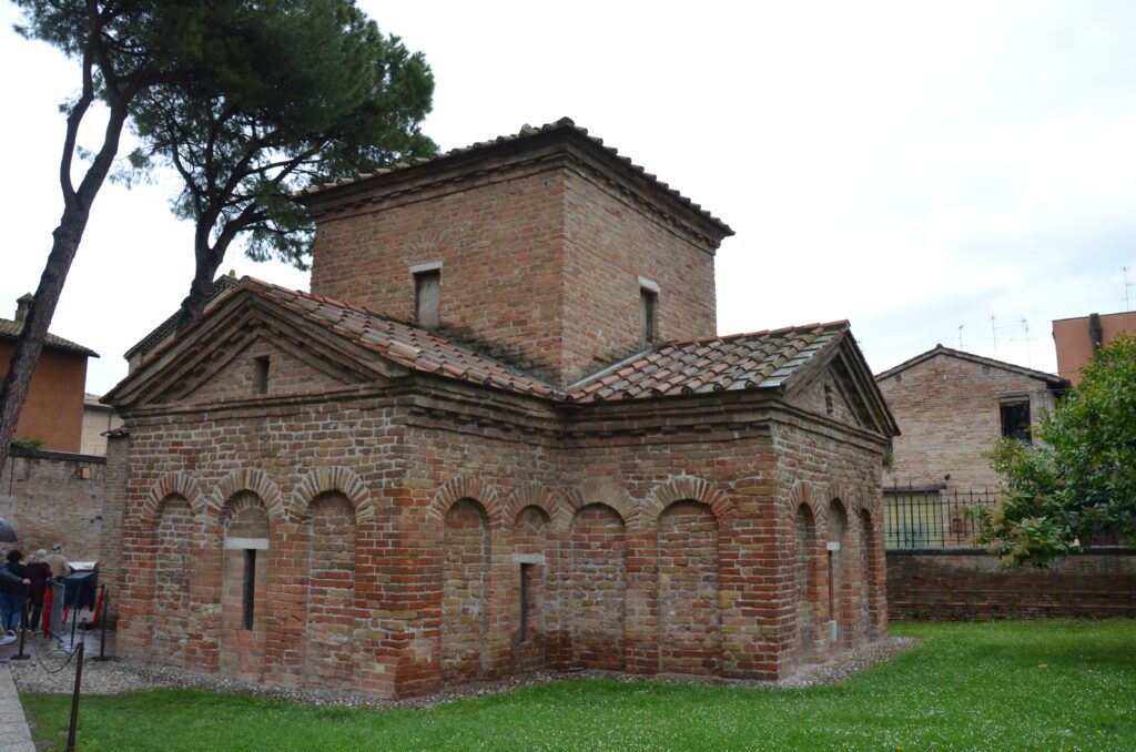 A lively street scene in an Italian town with people walking past red-brick and pastel-colored buildings lined with shops and cafes. Some people are chatting or taking photos, and flags are visible in the background near an arched gateway.