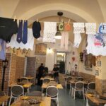 A restaurant with wooden tables and brown chairs on which people sit, clothes hanging on lines above their heads and between the walls.