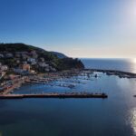 On the shore of the coast, on green hills, there is a sea with small houses, in the very blue sea there are piers and small boats.