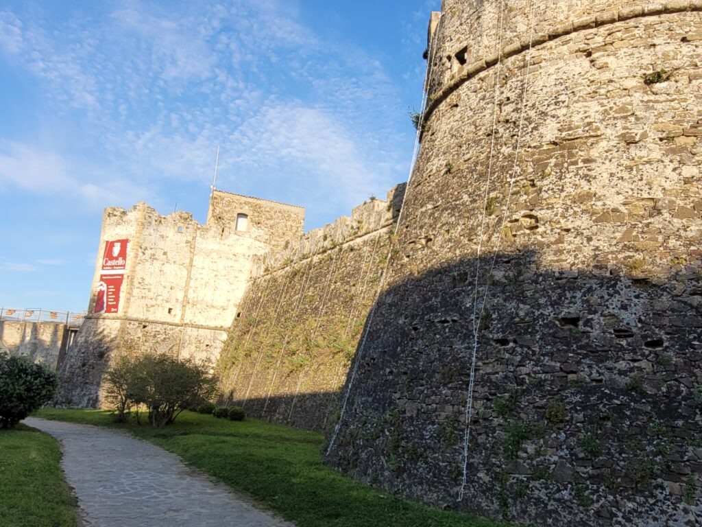 Exterior view of the imposing, rugged stone fortifications and tower of Agropoli's Aragonese Castle (Castello Angioino Aragonese). The defensive walls drop into shadow while a grassy path runs alongside them under a bright sky. The castle looks solid from the outside.
