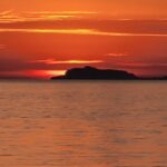 Intensywny, pomarańczowo-czerwony zachód słońca nad morzem. Ciemna sylwetka górzystej wyspy (prawdopodobnie Capri lub Ischia) odcina się na horyzoncie na tle nieba i odbicia światła na wodzie.