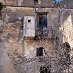 Abandoned stone house with cracked walls, a small balcony, and a leafless tree in front.