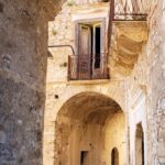 Stone archway leading to an old courtyard with weathered walls, a wooden door on a small balcony, and a metal gate blocking the entrance.