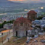 on the hill stands a small church with a red roof in the shape of a dome, behind it you can see the hills below and in the rays of the setting sun the city, in which a few lights shine