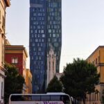 a tall, blue, narrow building stands in the city, in front of it there is a bus