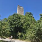 Ruined stone tower rising above trees on a hill, with two people walking along a country road in the foreground under a clear blue sky.