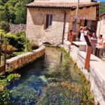 Clear stream running alongside an old stone building in a small Italian village, with people gathered near a terrace under the sun.