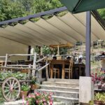 Outdoor seating area of a rustic Italian trattoria with wooden tables under a canopy, surrounded by flowers and a wooden cartwheel decoration.