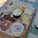 Wooden tray with assorted cheeses, cured meats, yogurt cups, and a beer bottle on a checkered tablecloth.