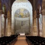 A view down the central aisle toward the apse of a Romanesque church interior. It shows stone columns with classical capitals and large stone arches supporting the nave. A terracotta-tiled path leads between rows of dark metal chairs towards a simple modern altar. The apse features a painted fresco under a half-dome ceiling. A piano is visible to the left in Lucca