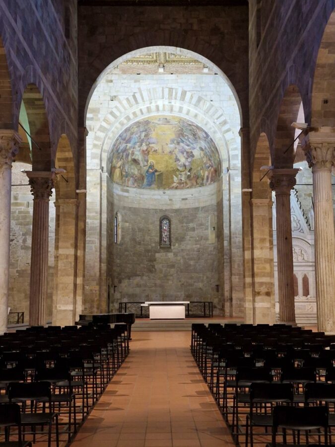 A view down the central aisle toward the apse of a Romanesque church interior. It shows stone columns with classical capitals and large stone arches supporting the nave. A terracotta-tiled path leads between rows of dark metal chairs towards a simple modern altar. The apse features a painted fresco under a half-dome ceiling. A piano is visible to the left in Lucca