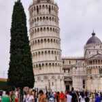 A tall, white leaning tower stands against the background of the cathedral, on one side there is a tall tree, and behind the tower there is a crowd of people spending a weekend break in Tuscany