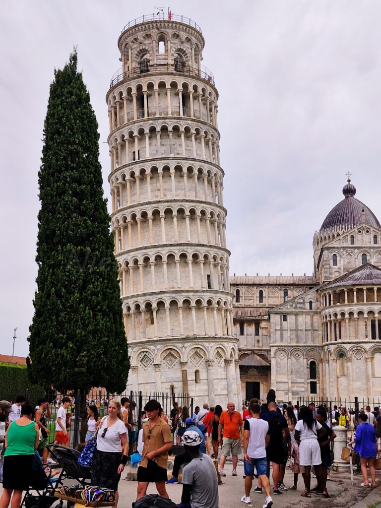 A tall, white leaning tower stands against the background of the cathedral, on one side there is a tall tree, and behind the tower there is a crowd of people.