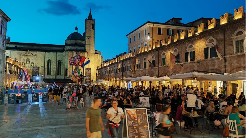 A bustling main square in Ascoli Piceno at dusk, featuring crowded outdoor restaurant seating under white awnings along historic arcaded buildings with warm lighting and crenellated rooflines. In the background, a large stone church with a dome and tower stands against a twilight sky, while people stroll across the polished travertine pavement of the piazza.