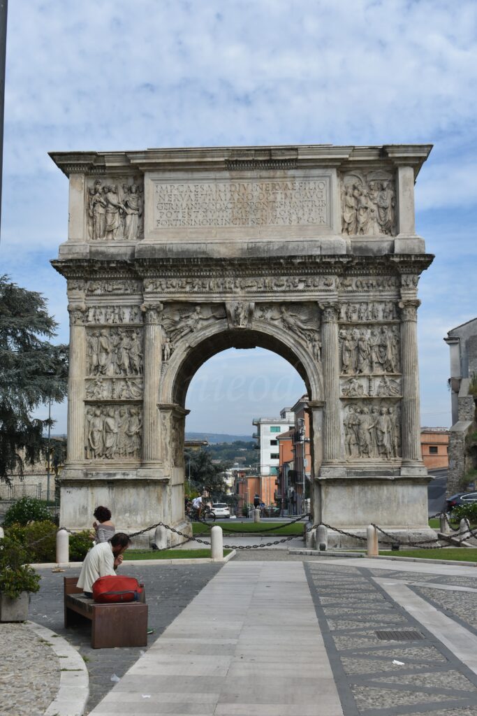 A tall ancient triumphal arch stands in the street, and in front of it a man sits on a bench.