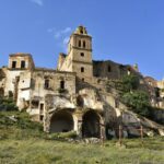 Ruined stone buildings on a hillside with a tall bell tower and arched structures, partly overgrown with vegetation.