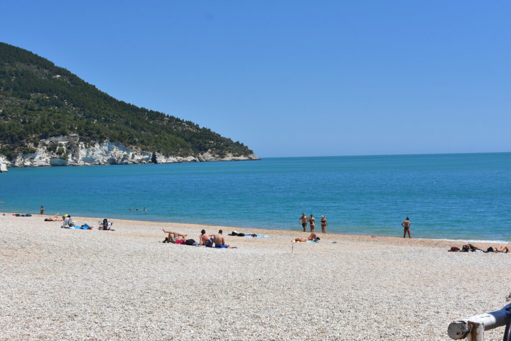 Beach with small pebbles, people sitting, a few people standing on the sea shore. In the background you can see a mountain covered with trees.