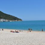 Beach with small pebbles, people sitting, a few people standing on the sea shore. In the background you can see a mountain covered with trees.