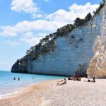 on a pebble Vignanotica Beach under a white cliff covered with trees, several people are sitting on a blanket, three more people are swimming in the blue sea, only their heads are visible
