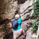 a blonde woman in a blue t-shirt and beige shorts has one leg on a rope looking from a high rock, and the other leg is on the ground