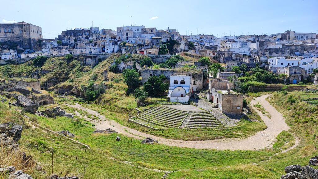 Old stone buildings and houses built into a rocky hillside, with patches of greenery and a dirt path leading toward them under a clear blue sky.
