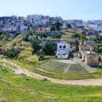 Old stone buildings and houses built into a rocky hillside, with patches of greenery and a dirt path leading toward them under a clear blue sky.