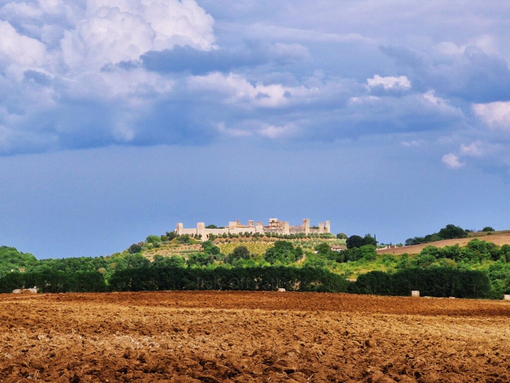 A distant view of the medieval walled town of Monteriggioni situated on a green hilltop in Tuscany. In the foreground is a wide, plowed field of dark brown soil, set under a dramatic sky with heavy, low-hanging white and grey clouds.