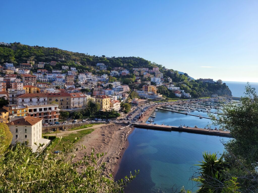is Agropoli worth visiting. View of Agropoli on the Cilento Coast in Italy, with colorful hillside houses overlooking the marina, beach, and calm Tyrrhenian Sea on a clear sunny day.