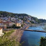 is Agropoli worth visiting. View of Agropoli on the Cilento Coast in Italy, with colorful hillside houses overlooking the marina, beach, and calm Tyrrhenian Sea on a clear sunny day.