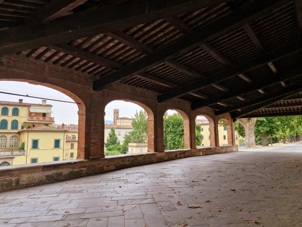 A view looking out from beneath a long, covered walkway with a dark wooden beam ceiling and brick archways. The stone-paved floor has scattered dry leaves. Through the open arches, yellow residential buildings, green trees, and a distant church tower are visible under an overcast sky.