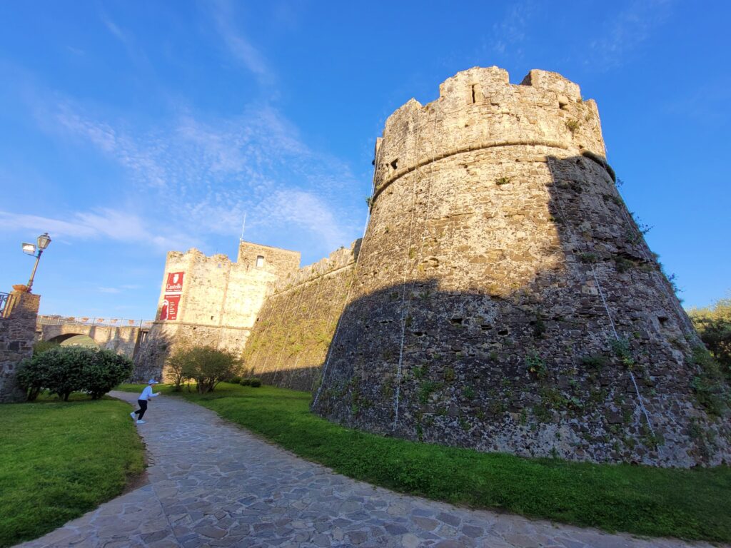 Exterior view of the imposing, rugged stone fortifications and tower of Agropoli's Aragonese Castle (Castello Angioino Aragonese). The defensive walls drop into shadow while a grassy path runs alongside them under a bright sky. The castle looks solid from the outside.