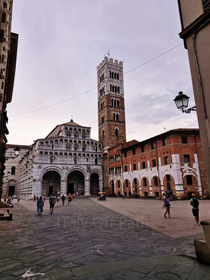 A wide shot of San Martino Cathedral in Lucca, Italy, featuring its white and grey marble facade with multiple tiers of open loggias and a tall, square stone bell tower. Pedestrians cross the paved piazza in front of the cathedral and an adjacent historic brick building on the right under a cloudy sky.
