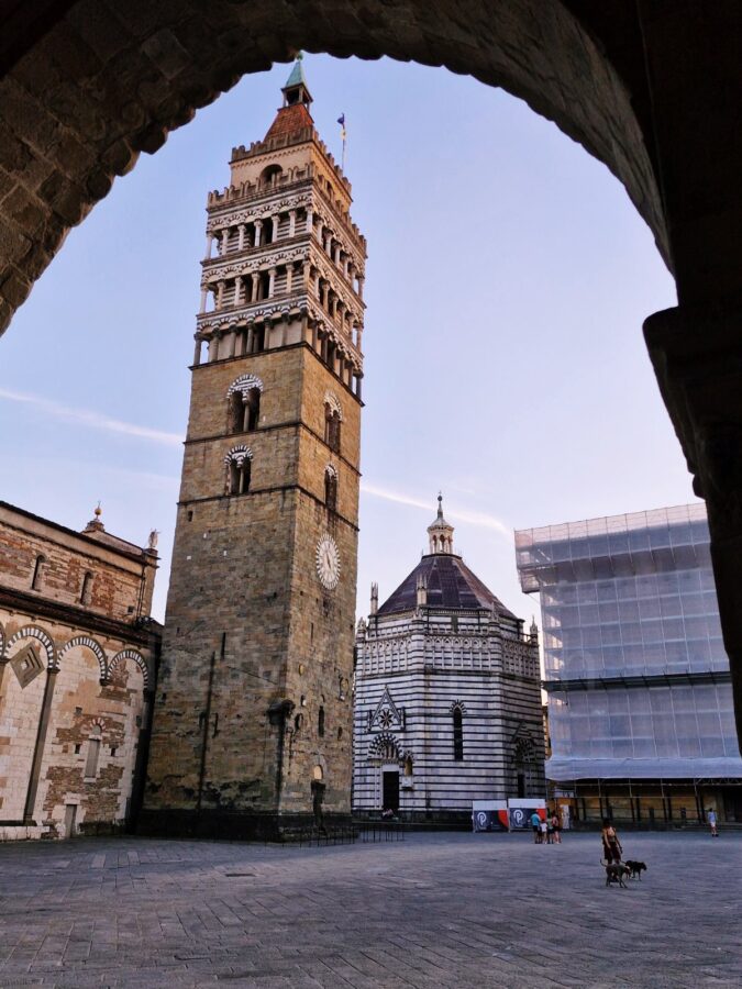 Widok kadrowany przez ciemny, kamienny łuk na Piazza del Duomo w Pistoi we Włoszech. Po lewej stronie wznosi się wysoka, kamienna dzwonnica katedry św. Zenona, obok której stoi pasiaste, marmurowe Baptysterium św. Jana. Przez szeroki, brukowany plac pod bezchmurnym, popołudniowym niebem spaceruje kilka osób, w tym jedna wyprowadzająca dwa psy.