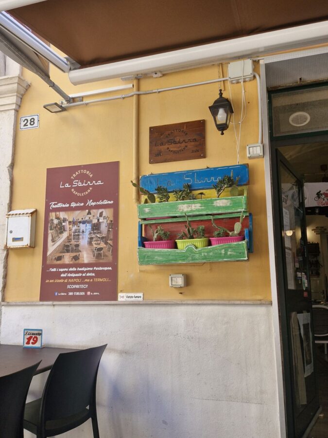 The entrance to Trattoria La Sbirra in Termoli, Molise, featuring a yellow wall, a decorative wooden planter with cacti, and a promotional sign for the restaurant.