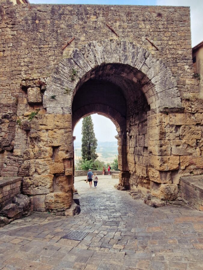 A massive, ancient Etruscan stone archway in Volterra, Tuscany, constructed from large, weathered block masonry. Through the deep arch, people walk along a paved path, leading to a view of a single tall cypress tree and distant, hazy Tuscan hills.