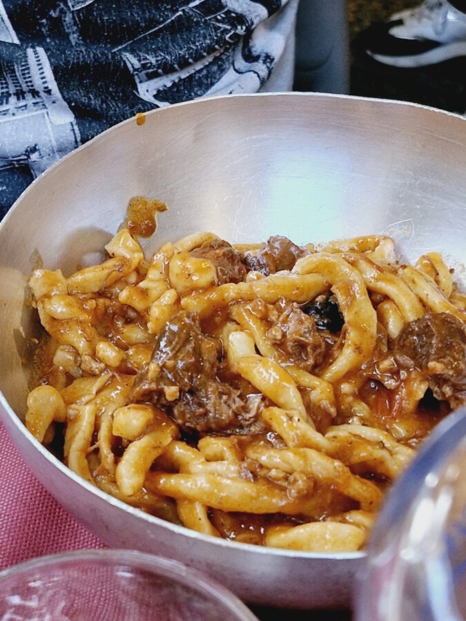 A close-up of thick, hand-rolled Tuscan pasta coated in a rich, chunky meat ragu, served in a deep metal bowl.