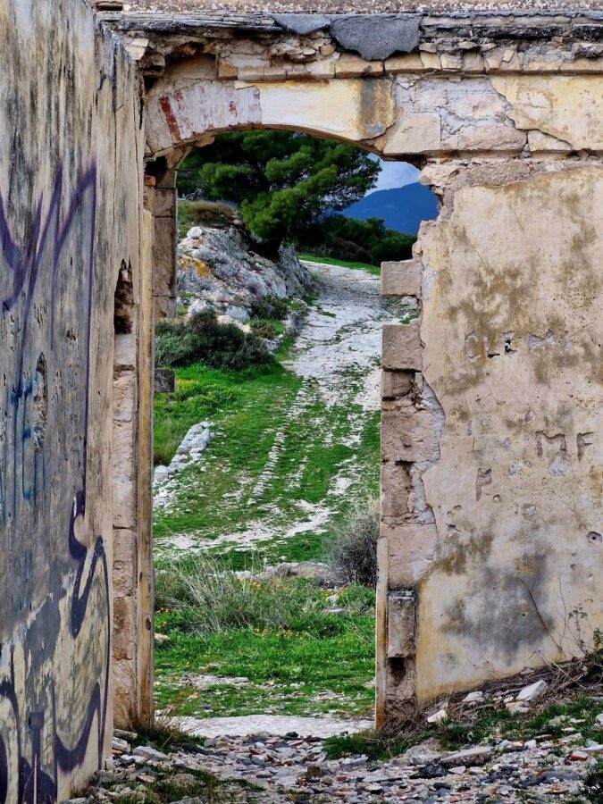 View through a cracked, crumbling plaster doorway of a ruined stone building covered in graffiti. The opening frames a grassy path leading past rocks and a green pine tree under a cloudy sky.
