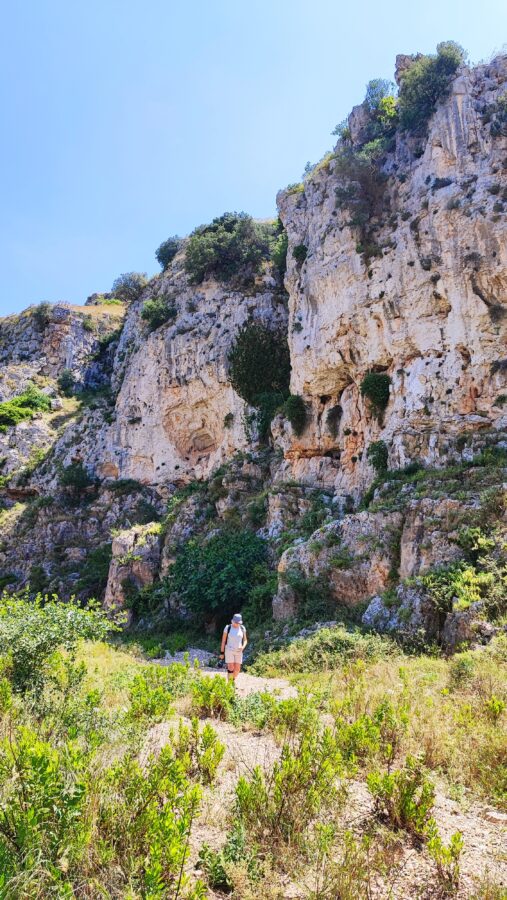 Hiker walking along a trail at the base of towering limestone rock cliffs in Gravina di Ginosa, Puglia.
