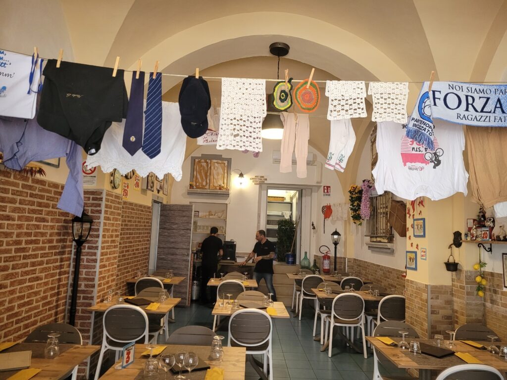 The interior of Trattoria La Sbirra in Termoli, Molise, featuring dining tables, brick-accented walls, an arched ceiling, and clotheslines strung across the room hanging shirts, ties, crocheted cloths, and sports scarves.