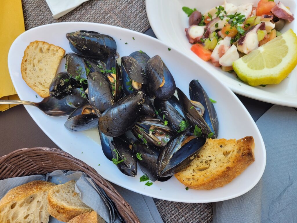 An oval white plate filled with steamed mussels in a light broth, garnished with fresh parsley and served with toasted bread slices.