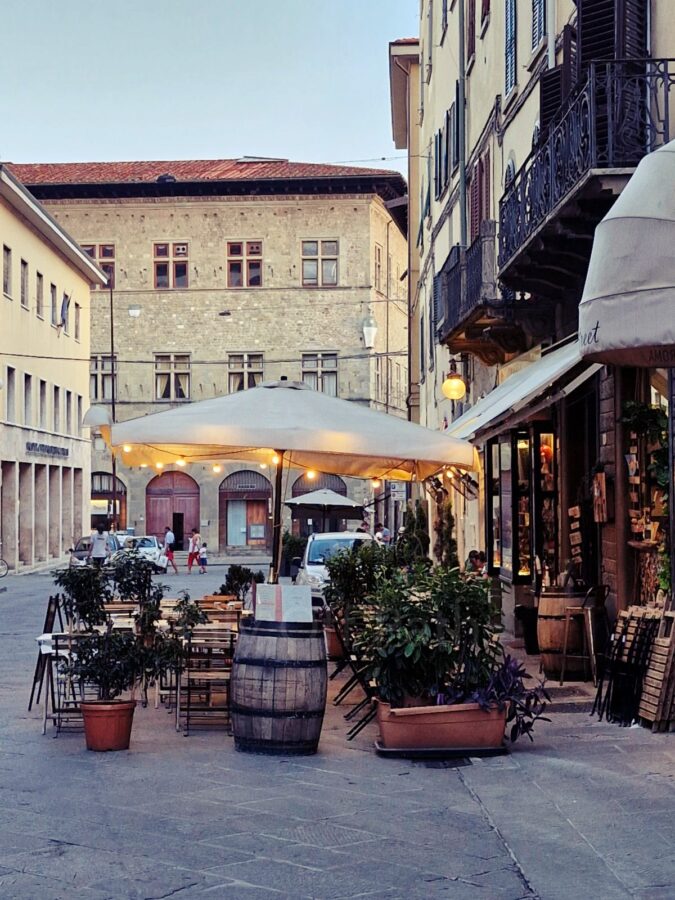 Ogródek restauracyjny na brukowanej ulicy w Pistoi we Włoszech. Drewniane stoliki i krzesła są ustawione pod dużym parasolem z rozwieszonymi lampkami, obok ozdobnej drewnianej beczki po winie. Ulicę otaczają zabytkowe kamienice z okiennicami, a w tle widać spacerujących przechodniów.