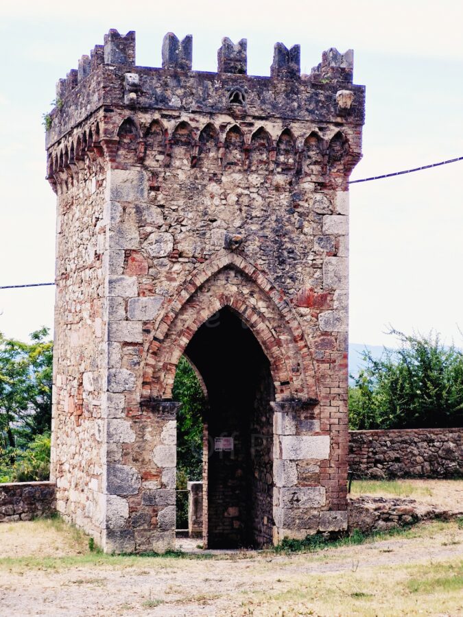 An ancient stone and brick gate tower with medieval features in Frosini, Tuscany, featuring a crenellated top and a Gothic pointed archway.