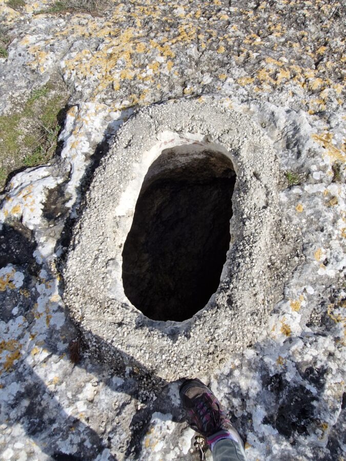 An oval opening carved into a rocky, textured ground, leading into a dark, deep hole of an ancient tomb in Mattinata. The opening has a raised, rough, light-colored concrete or stone border. The surrounding rock surface is uneven and covered with patches of yellow and white lichen. A person's foot wearing a hiking shoe is visible at the bottom of the frame, providing scale.